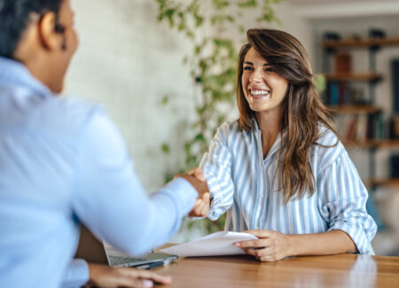 Eine lächelnde Frau in einem gestreiften Hemd sitzt an einem Schreibtisch, hält Papiere in der Hand und schüttelt einer anderen Person in einem Büro gegenüber die Hand.