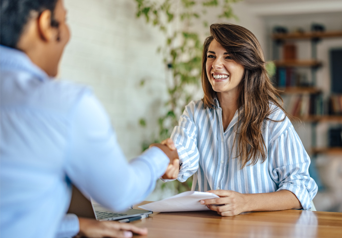 Eine lächelnde Frau in einem gestreiften Hemd sitzt an einem Schreibtisch, hält Papiere in der Hand und schüttelt einer anderen Person in einem Büro gegenüber die Hand.