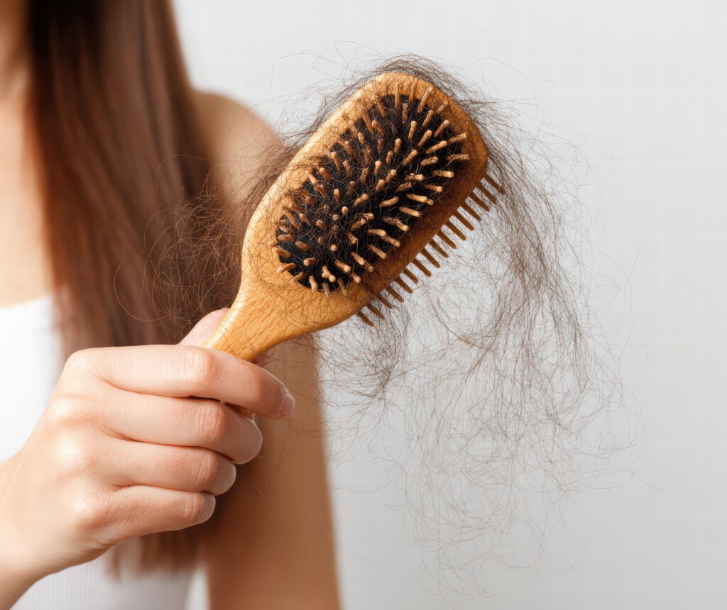 Eine Person mit langem braunem Haar hält eine hölzerne Haarbürste, die mit verhedderten Haarsträhnen gefüllt ist, was auf Haarausfall hinweist.