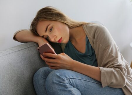 Eine junge Frau mit langen blonden Haaren sitzt auf einer grauen Couch, stützt den Kopf auf ihren Arm und schaut nachdenklich auf ihr Smartphone. Sie wirkt nachdenklich oder besorgt und trägt legere Kleidung in einer entspannten Umgebung.
