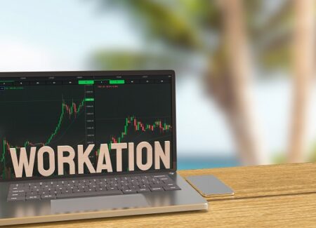 Ein Laptop mit Aktiencharts und dem Wort WORKATION auf dem Bildschirm steht auf einem Tisch im Freien