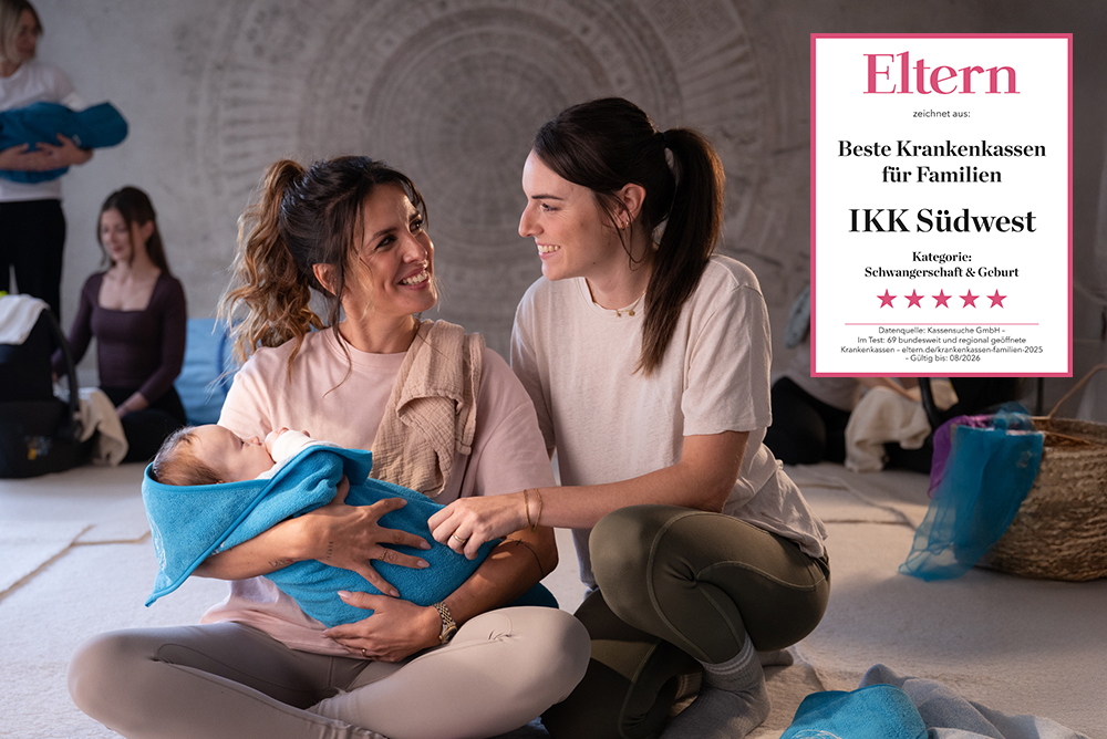 Zwei Mütter sitzen in einem Eltern-Baby-Kurs auf dem Boden, unterhalten sich und lächeln; eine hält ein in eine blaue Decke gewickeltes Baby. Im Hintergrund sind weitere Eltern und Babys zu sehen. Im Vordergrund ist eine Plakette der IKK Südwest zu sehen.