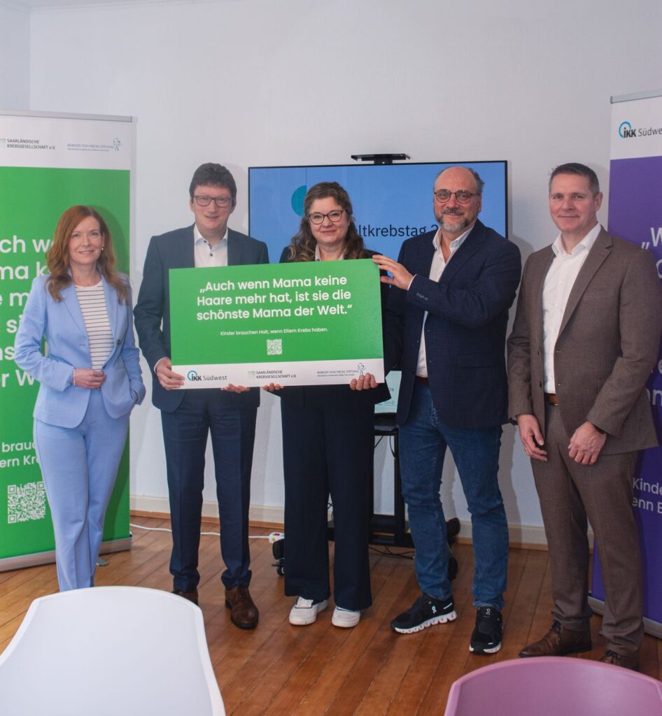 Fünf Personen in Geschäftskleidung halten ein grün-weißes Schild mit dem Text: "Auch wenn Mama keine Haare mehr hat, ist sie die schönste Mama der Welt."