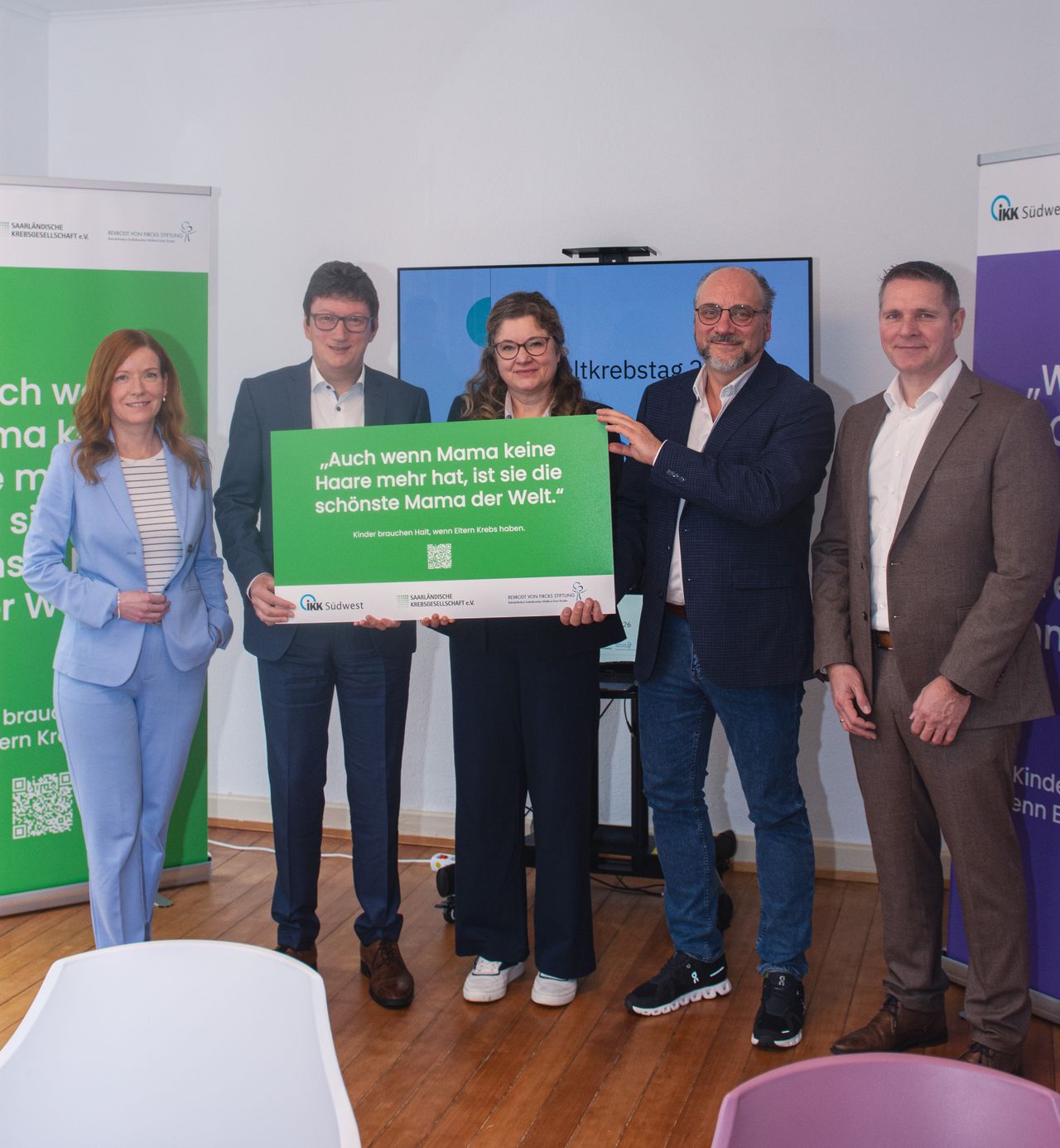 Fünf Personen in Geschäftskleidung halten ein grün-weißes Schild mit dem Text: "Auch wenn Mama keine Haare mehr hat, ist sie die schönste Mama der Welt."