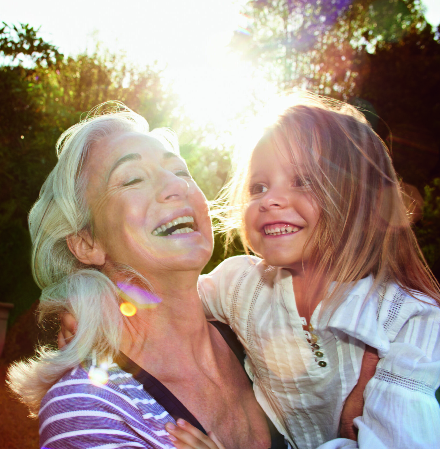 Eine ältere Frau mit grauem Haar lächelt und hält ein junges Mädchen mit langen Haaren im Arm. Sie sind draußen in einem sonnigen, grünen Garten, beide lachen fröhlich im Sonnenlicht, das hinter ihnen scheint.