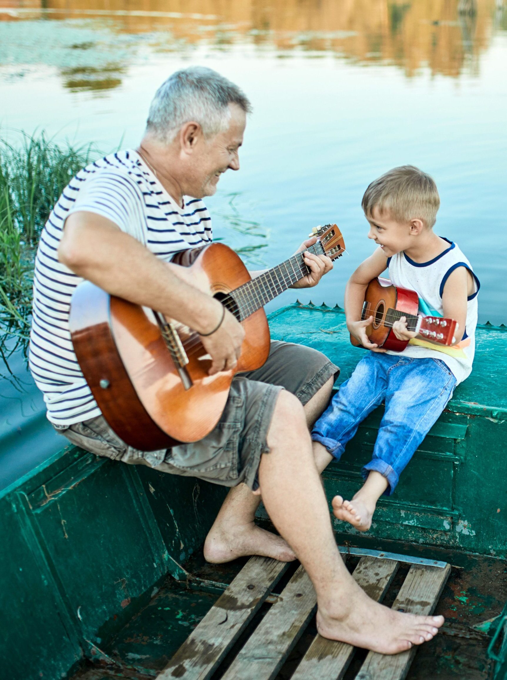 Ein älterer Mann und ein kleiner Junge sitzen barfuß auf einem grünen Boot, lächeln und spielen gemeinsam Akustikgitarre auf einem ruhigen See, umgeben von der Natur.