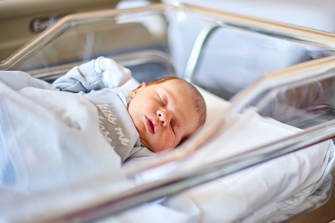 Ein neugeborenes Baby, eingewickelt in eine hellblaue Decke, schl&auml;ft friedlich in einem Krankenhausbettchen.