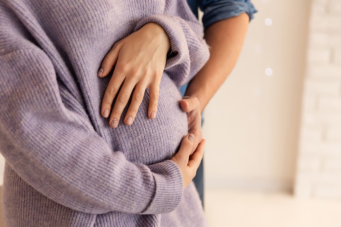 Nahaufnahme einer schwangeren Person, die einen lavendelfarbenen Pullover trägt und deren Hände sanft auf ihrem Bauch ruhen. Eine andere Person steht dahinter und legt eine stützende Hand auf die Seite der Schwangeren.