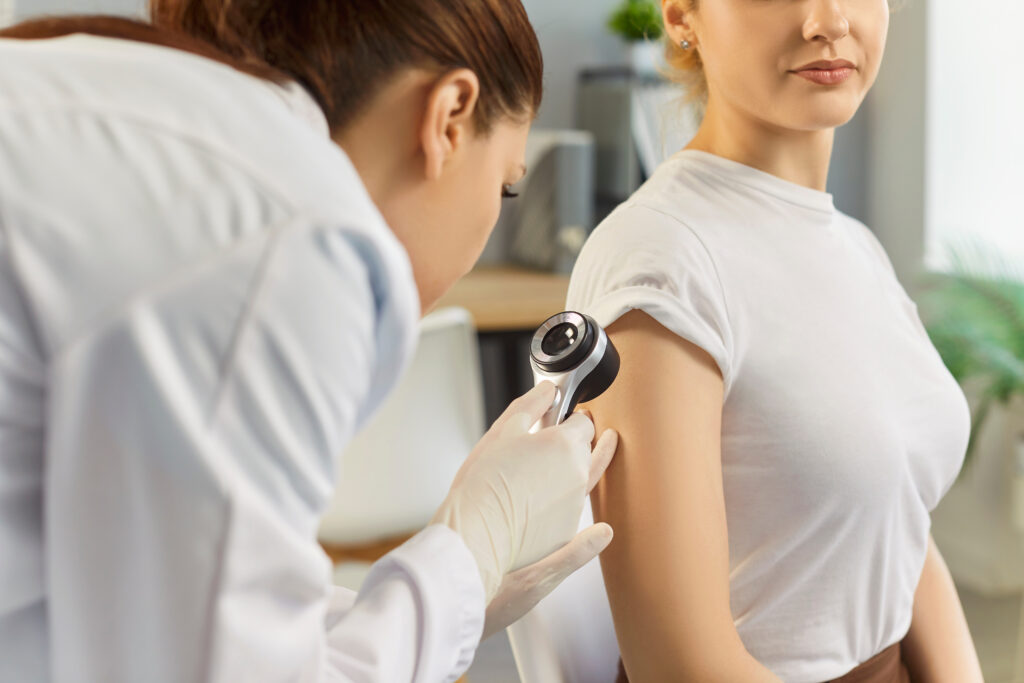 Eine Ärztin mit Handschuhen untersucht in einer Arztpraxis den Oberarm einer Frau mit einem Dermatoskop.