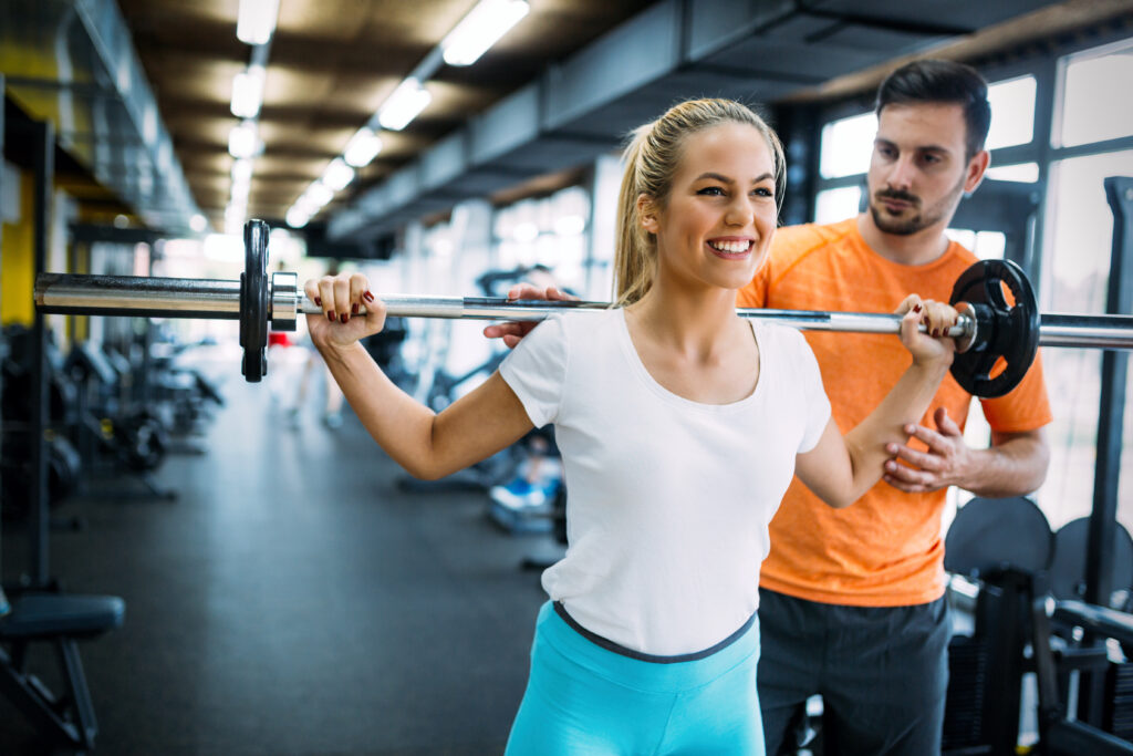 Eine Frau in Sportkleidung hebt in einem Fitnessstudio eine Langhantel auf ihre Schultern und lächelt, während ein Trainer hinter ihr steht und ihre Bewegung korrigiert.