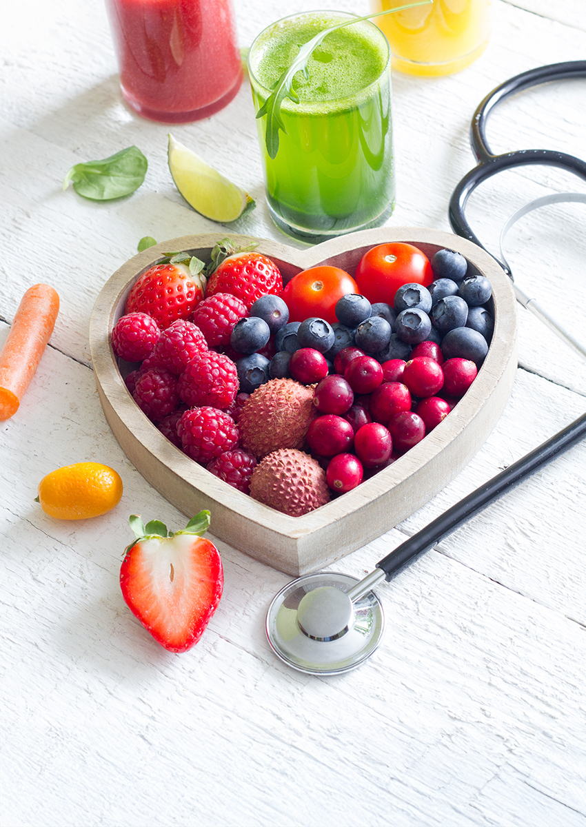 Eine herzförmige Schale, gefüllt mit verschiedenen Beeren, Litschi, Kirschtomaten und Preiselbeeren, steht neben einem Stethoskop und drei Gläsern mit bunten Säften auf einer weißen Holzfläche.
