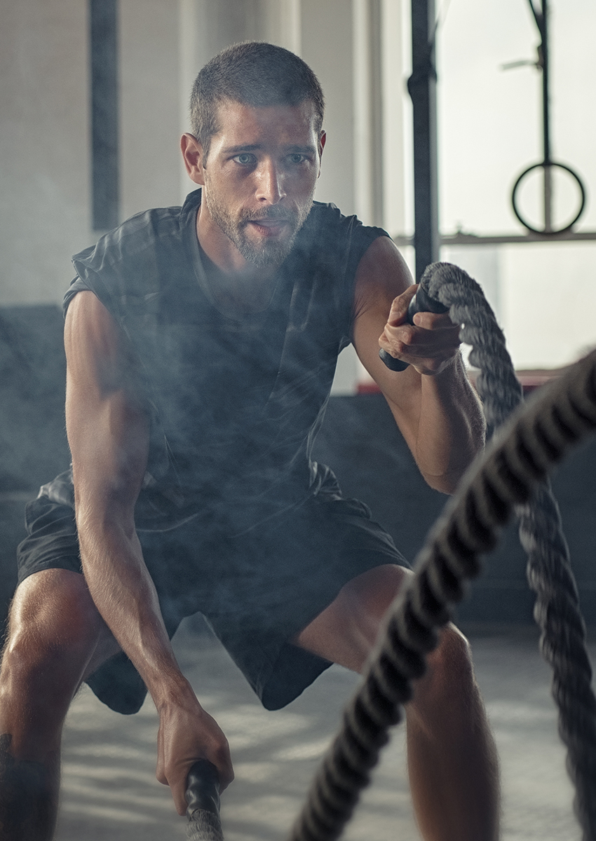 Ein Mann mit kurzen Haaren und Bart trainiert intensiv mit Kampfseilen in einem Fitnessstudio, er trägt ein ärmelloses schwarzes Hemd und kurze Hosen. Das Sonnenlicht fällt durch große Fenster hinter ihm.