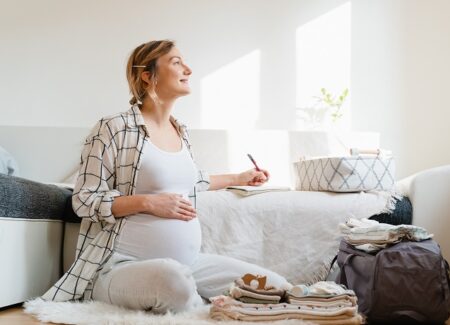 Eine schwangere Frau sitzt in einem Zimmer auf dem Boden. Sie schreibt in ein Notizbuch, vor ihr eine gepackte Krankenhaustasche.