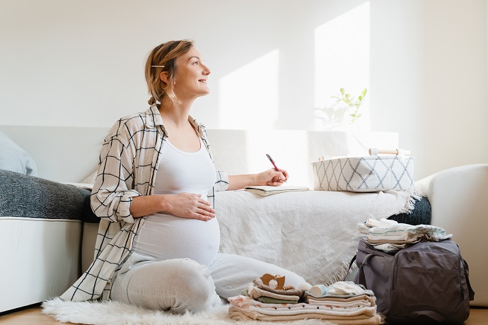 Eine schwangere Frau sitzt in einem Zimmer auf dem Boden. Sie schreibt in ein Notizbuch, vor ihr eine gepackte Krankenhaustasche.