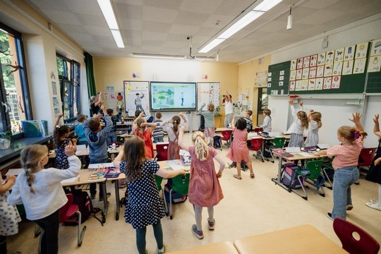 In einem Klassenzimmer stehen die Kinder neben ihren Tischen, heben die Arme und nehmen an einer interaktiven Lektion teil, die auf einem Bildschirm an der Stirnseite gezeigt wird, während ein Lehrer die Aktivität leitet.