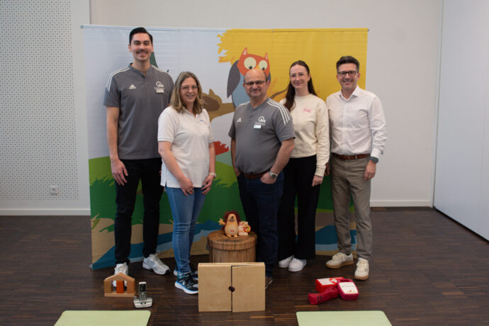 Fünf Erwachsene stehen in einem Raum vor einem bunten Hintergrund mit einer illustrierten Eule. Auf dem Boden vor ihnen befinden sich pädagogische Spielzeuge und ein Erste-Hilfe-Equipment.