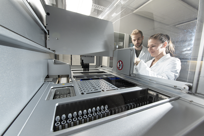 Zwei Personen in Laborkitteln bedienen eine große, hochmoderne Labormaschine, die mit Reagenzgläsern und Fläschchen gefüllt ist.