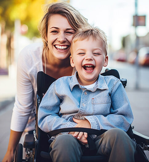 Ein kleiner Junge im Rollstuhl und eine Frau, beide lachend, befinden sich im Freien auf einem städtischen Bürgersteig mit verschwommenen Gebäuden und Autos im Hintergrund.