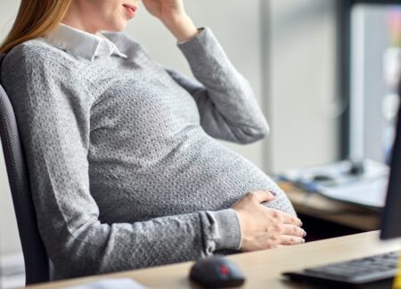 Eine schwangere Frau sitzt an einem Schreibtisch. Sie stützt eine Hand auf ihren Bauch.