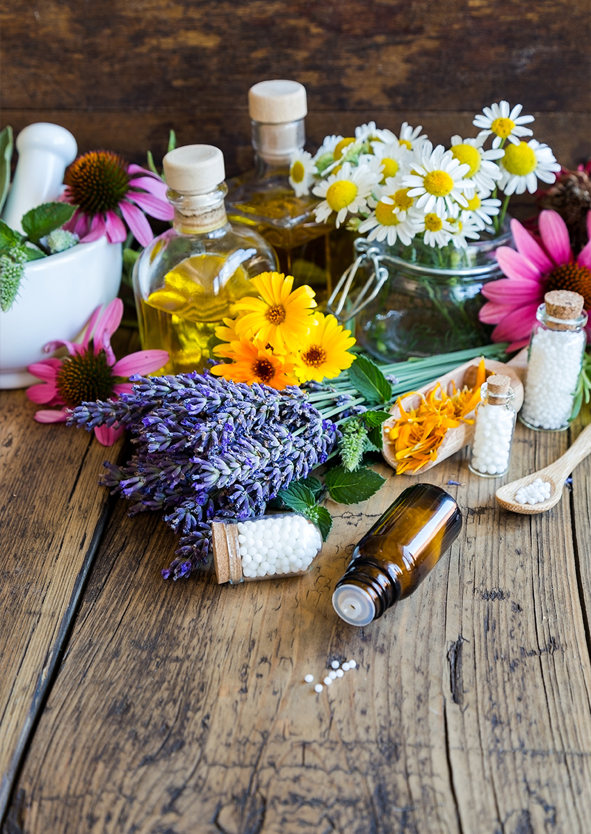 Auf einer rustikalen Holzfläche sind verschiedene frische Blumen, Ölflaschen, ein Glasgefäß, ein braunes Medizinfläschchen, weiße homöopathische Pillen und ein Mörser und Stößel angeordnet.