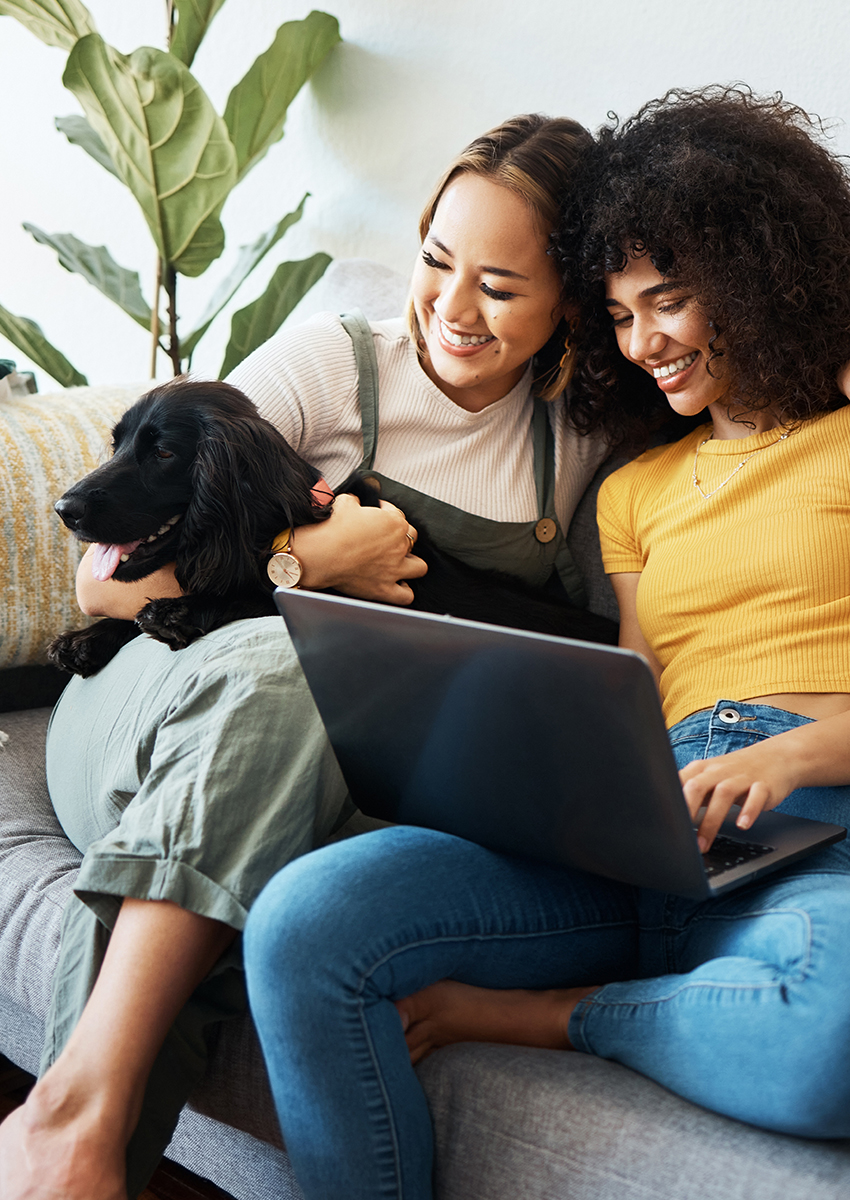Zwei Frauen sitzen lächelnd auf einer Couch, eine hält einen schwarzen Hund, die andere arbeitet mit einem Laptop. Sie wirken glücklich und entspannt, mit einer Grünpflanze im Hintergrund.