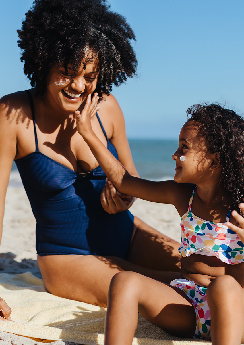 Eine Frau in einem marineblauen Badeanzug und ein junges Mädchen in einem bunt gemusterten Badeanzug lächeln und spielen zusammen am Strand, wobei sie sich gegenseitig mit Sonnencreme eincremen, während im Hintergrund das Meer und der blaue Himmel zu sehen sind.