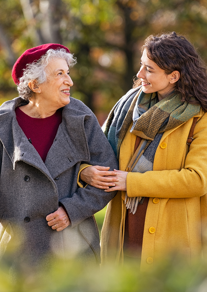 Eine ältere Frau in einem grauen Mantel und einer roten Baskenmütze lächelt, während sie Arm in Arm mit einer jüngeren Frau in einem gelben Mantel und einem Schal spazieren geht, die beide einen schönen Tag im Freien genießen.