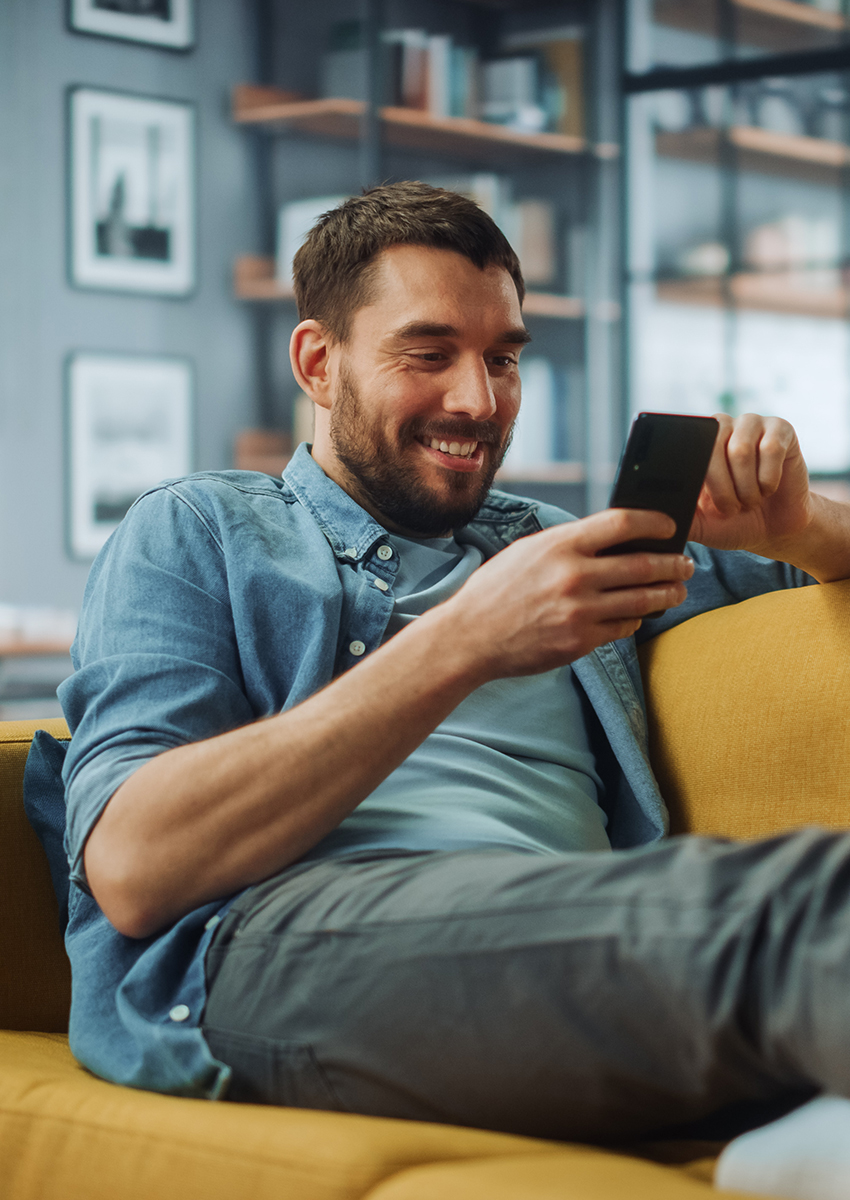 Ein Mann mit Bart, blauem Hemd und grauer Hose, sitzt auf einem gelben Sofa und schaut lächelnd auf sein Smartphone in einem modernen, gemütlichen Wohnzimmer mit Regalen und gerahmten Fotos an der Wand.