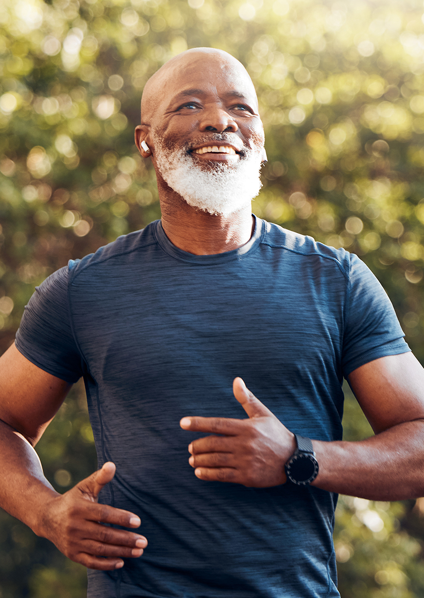 Ein lächelnder älterer Mann mit weißem Bart joggt im Freien, während er ein dunkles Sporthemd, eine Smartwatch und kabellose Ohrhörer trägt, mit Grünzeug im unscharfen Hintergrund.