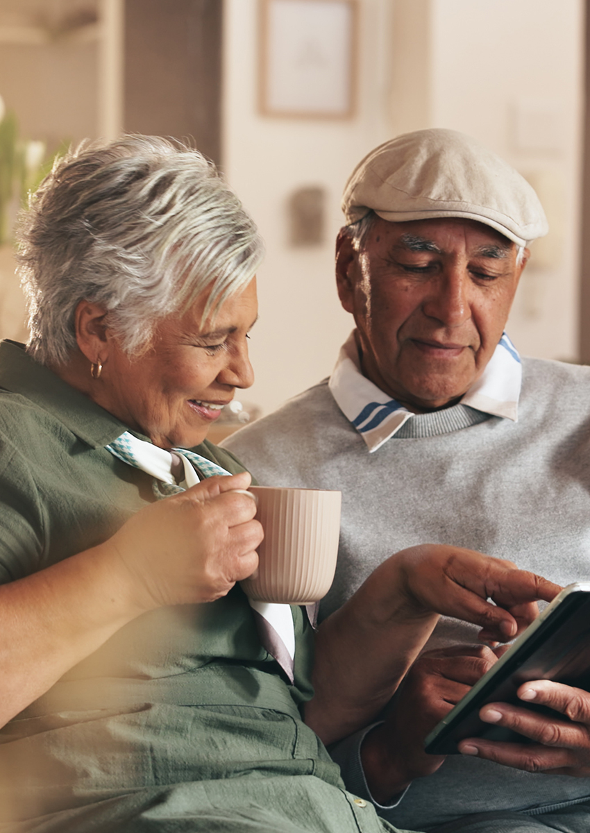 Ein älteres Paar sitzt eng beieinander auf einer Couch. Die Frau lächelt und hält eine Tasse in der Hand, während der Mann eine Mütze trägt und ein Tablet benutzt, beide scheinen glücklich und mit dem Gerät beschäftigt zu sein.