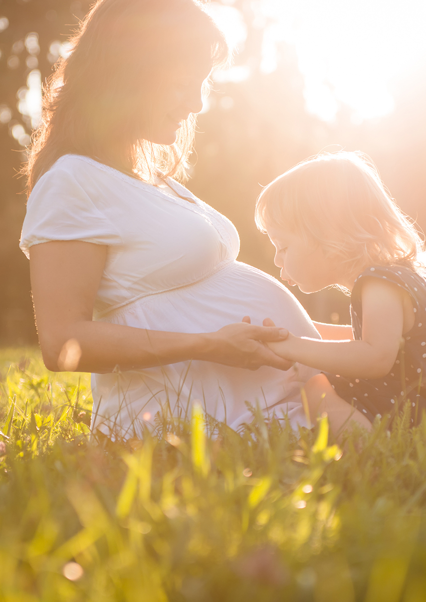 Ein kleines Kind küsst den Bauch einer schwangeren Frau, während sie gemeinsam im warmen Sonnenlicht im Gras sitzen.