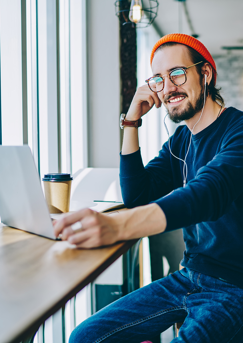 Ein lächelnder Mann mit Brille, leuchtend orangefarbener Mütze und legerer Kleidung sitzt an einem Holztisch am Fenster und arbeitet mit Kopfhörern an einem Laptop. Neben ihm steht ein Kaffeebecher zum Mitnehmen.