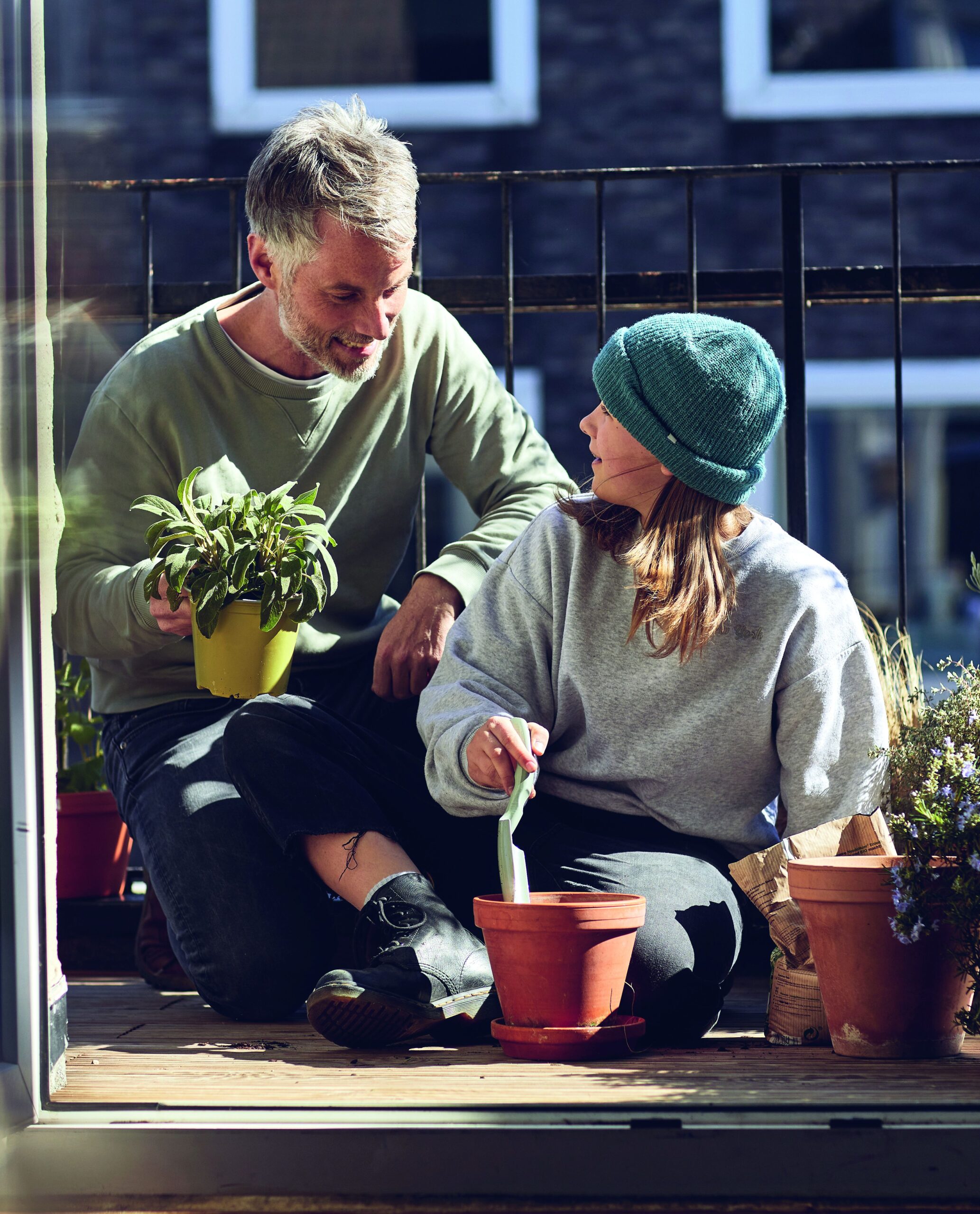 Ein Mann und ein Mädchen sitzen auf einem Balkon und gärtnern gemeinsam. Der Mann hält eine Topfpflanze, während das Mädchen, die eine grüne Mütze trägt, einen Tontopf vorbereitet. Das Sonnenlicht scheint auf sie, und die Pflanzen liegen verstreut herum.