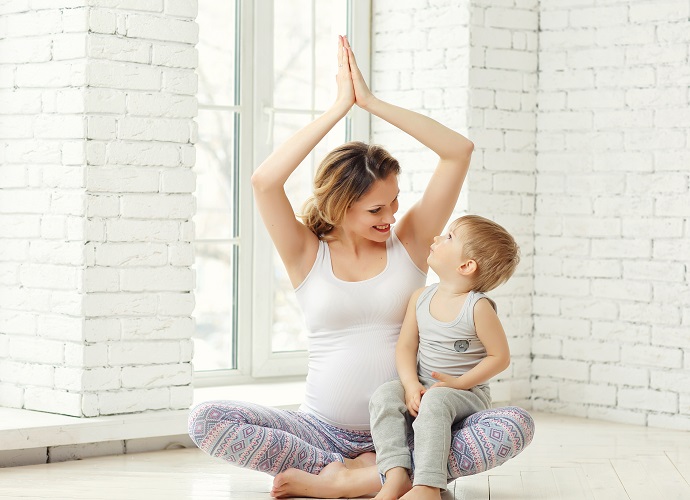 Eine schwangere Frau sitzt im Schneidersitz auf dem Boden und hält ihre Hände in einer Yoga-Pose über den Kopf. Ein kleines Kind sitzt auf ihrem Schoß und schaut ihr zu.