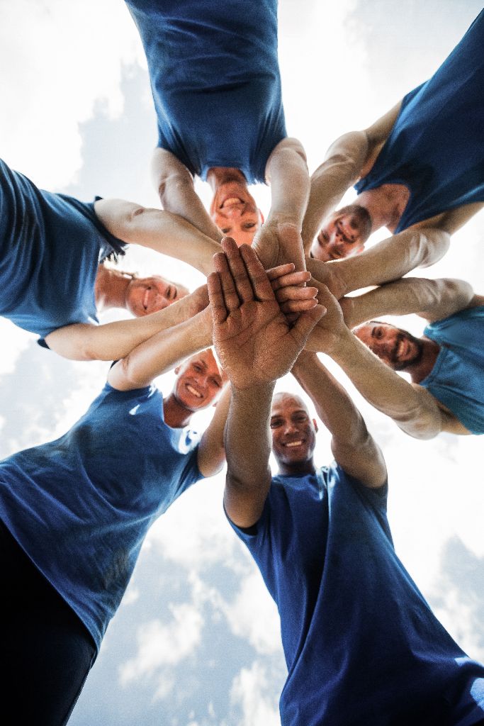 Sechs Personen in blauen Hemden stehen in einem Kreis, lächeln und legen ihre Hände in der Mitte zusammen, von unten gesehen gegen einen hellen, teilweise bewölkten Himmel.