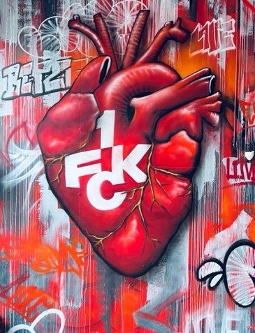 Ein Graffiti-Wandbild eines anatomischen Herzens mit dem Wappen des 1. FC Kaiserslautern darüber gemalt, umgeben von bunten roten, weißen und grauen Graffiti-Tags und aufgesprühten Texturen.