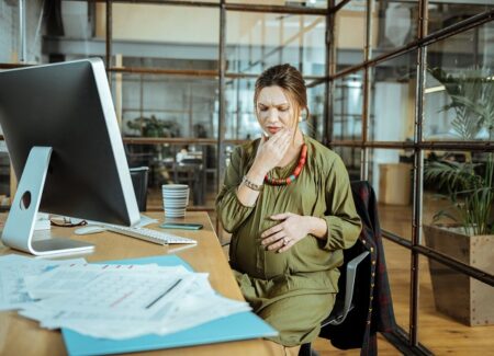 Eine schwangere Frau sitzt an einem Schreibtisch in einem B&uuml;ro, h&auml;lt sich mit einer Hand den Bauch und ber&uuml;hrt mit besorgter Miene ihr Kinn.