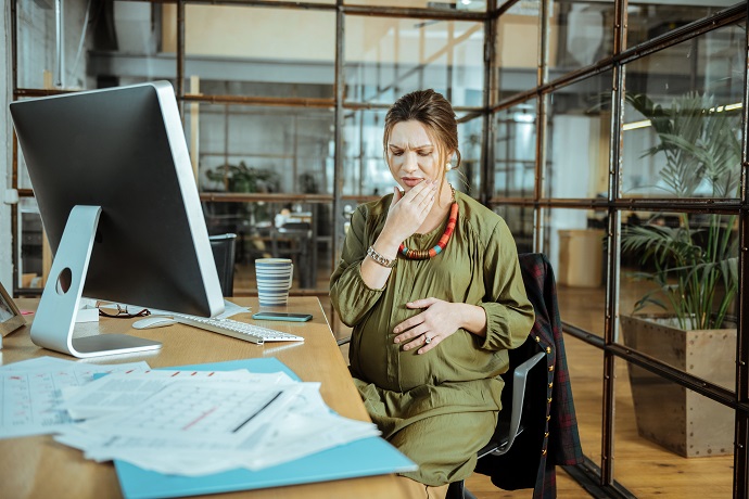 Eine schwangere Frau sitzt an einem Schreibtisch in einem Büro, hält sich mit einer Hand den Bauch und berührt mit besorgter Miene ihr Kinn.