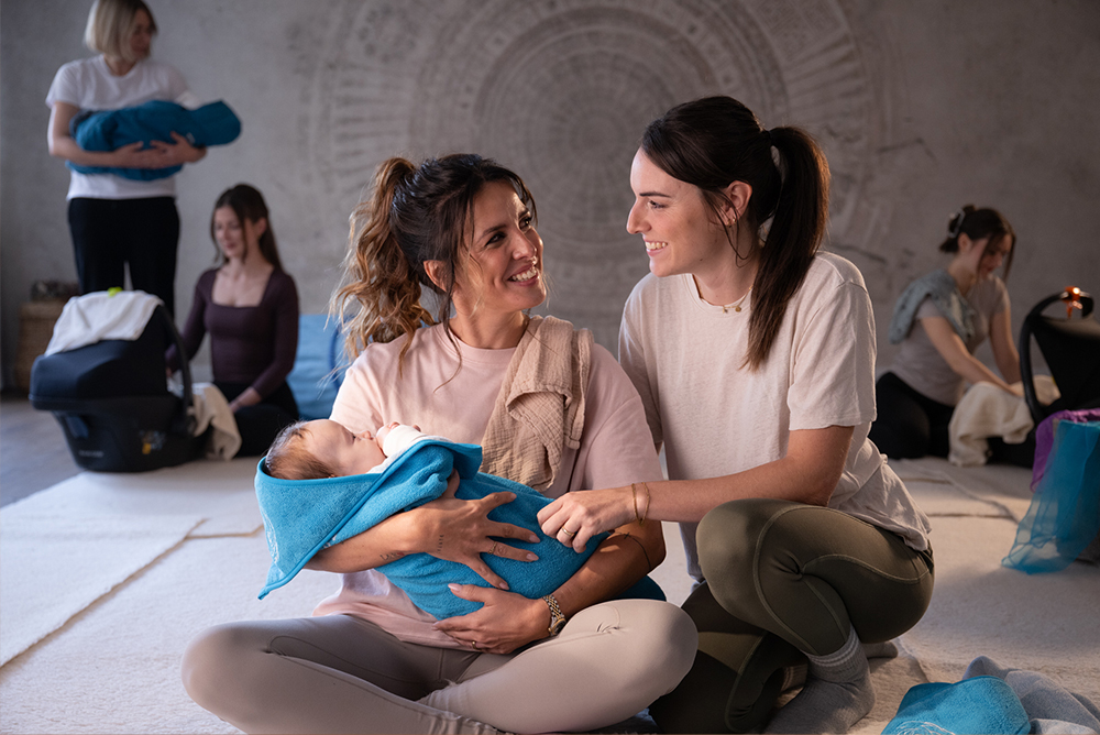 Zwei Frauen sitzen auf dem Boden und lächeln einander an; eine von ihnen hält ein Baby im Arm, das in eine blaue Decke gewickelt ist. Im Hintergrund sind weitere Frauen mit Babys in einem ruhigen, sanft beleuchteten Raum zu sehen.