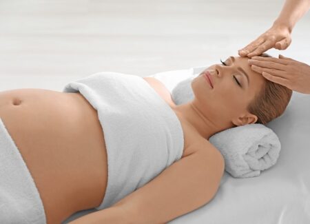 Eine schwangere Frau liegt auf einer Massageliege, ihr K&ouml;rper ist mit einem Handtuch bedeckt, sie hat die Augen geschlossen und erh&auml;lt von den H&auml;nden eines Therapeuten eine sanfte Stirnmassage.