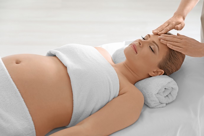 Eine schwangere Frau liegt auf einer Massageliege, ihr Körper ist mit einem Handtuch bedeckt, sie hat die Augen geschlossen und erhält von den Händen eines Therapeuten eine sanfte Stirnmassage.