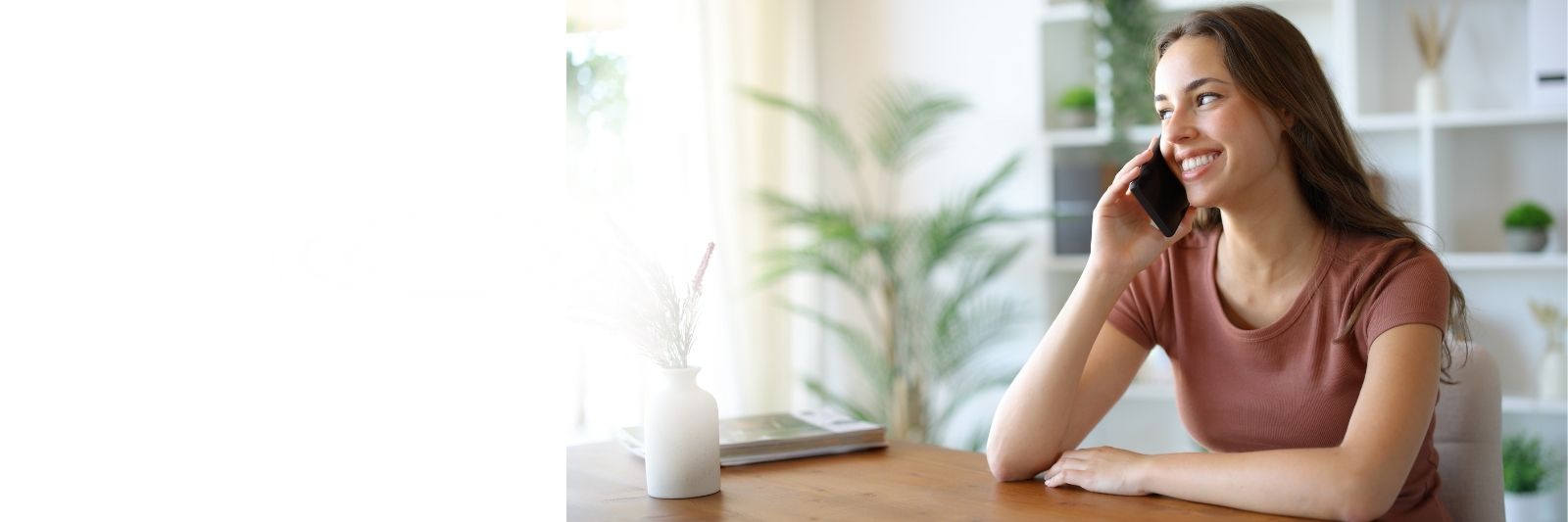 Eine Frau sitzt an einem Holztisch und l&auml;chelt, w&auml;hrend sie mit ihrem Smartphone telefoniert.