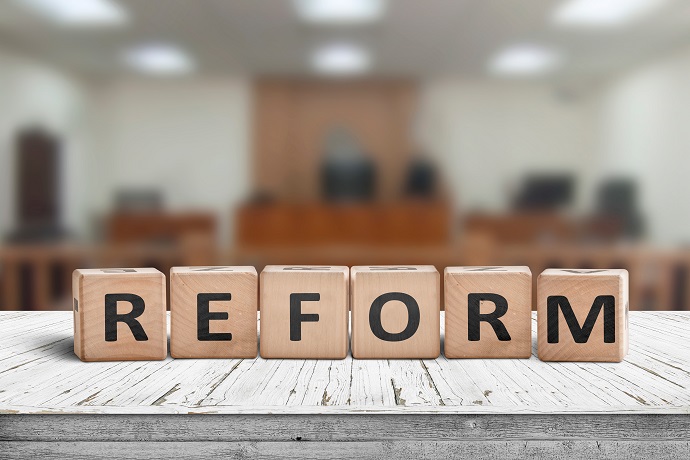 Holzklötze, die das Wort „REFORM“ bilden, liegen auf einem Tisch vor einem verschwommenen Hintergrund, der einen Gerichtssaal darstellt.