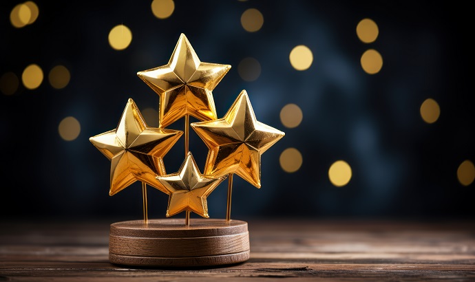 Auf einem Holztisch steht eine goldfarbene, sternförmige Trophäe mit fünf Sternen auf einem Holzsockel. Verschwommene goldene Lichter zieren den dunklen Hintergrund.