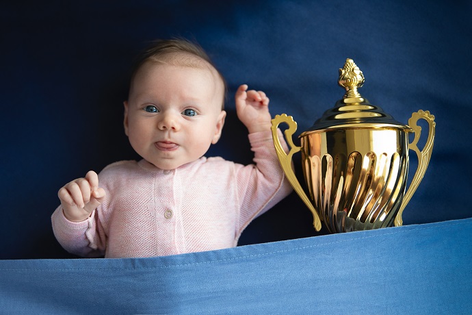 Ein Baby in einem rosa Strampler liegt auf einer blauen Decke neben einer großen goldenen Trophäe.