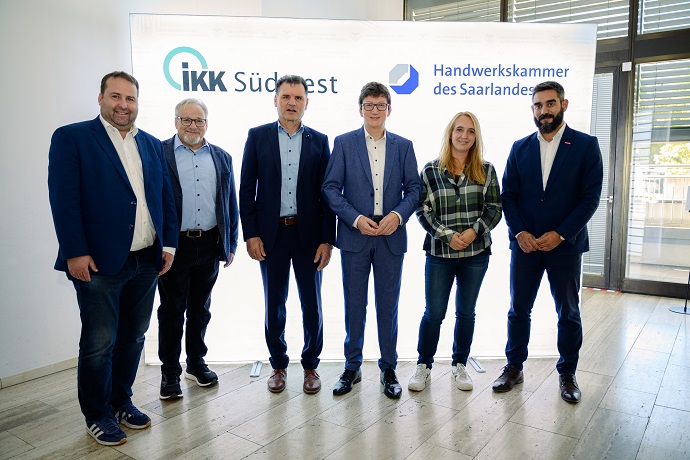 Sechs Personen stehen in einer Reihe, gekleidet in Business- oder Smart-Casual-Kleidung, vor einem wei&szlig;en Hintergrund mit den Logos des IKK S&uuml;dwest und der Handwerkskammer des Saarlandes.