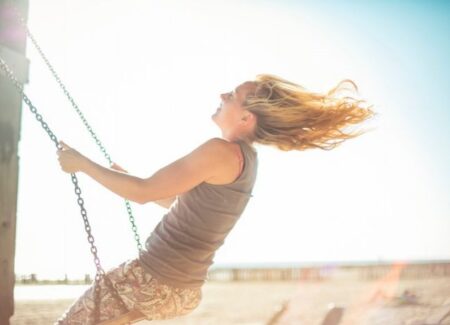 Eine Person mit langen Haaren schaukelt an einem strahlenden, sonnigen Tag im Freien, den Kopf nach hinten geneigt, w&auml;hrend ihr Haar im Wind weht.