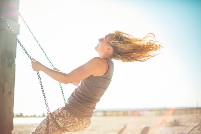 Eine Person mit langen Haaren schaukelt an einem strahlenden, sonnigen Tag im Freien, den Kopf nach hinten geneigt, während ihr Haar im Wind weht.