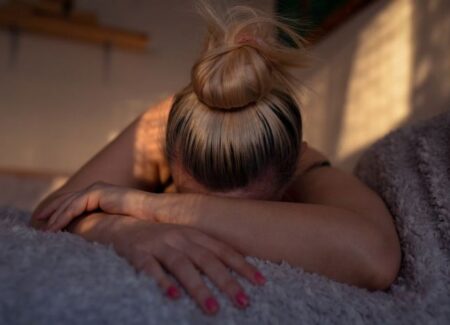 Eine Frau mit blonden Haaren, die zu einem Dutt zusammengebunden sind, liegt mit dem Gesicht nach unten auf einem Bett, die Arme vor dem Gesicht verschr&auml;nkt.