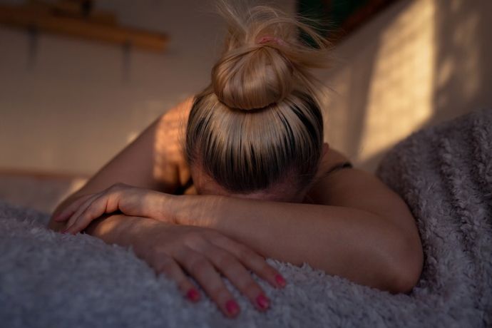 Eine Frau mit blonden Haaren, die zu einem Dutt zusammengebunden sind, liegt mit dem Gesicht nach unten auf einem Bett, die Arme vor dem Gesicht verschränkt.
